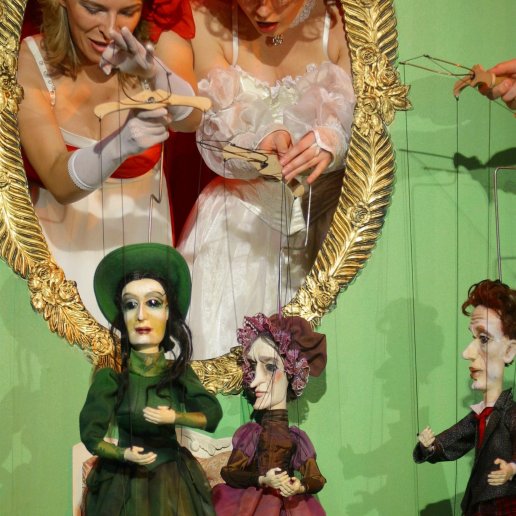 Dwie postaci w oknie dekoracyjnym, owalnym, ozdobionym na złoto, animują marionetki. Agnieszka w czerwonym kapeluszu, druga aktorka w czerwonej peruce. Obydwie ubrane na biało.