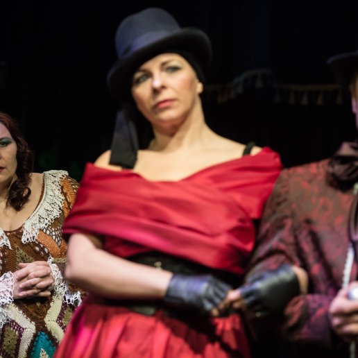 Troje aktorów, pośrodku Agnieszka elegancka w czerwonej sukni, czarnych rękawiczkach i czarnym nakryciu głowy. Trzyma pod rękę mężczyznę.