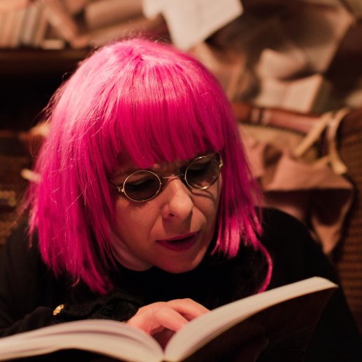 Agnieszka w różowej jaskrawej peruce z grzywką i okrągłych okularach na nosie. Twarz zatopiona w otwartej książce.