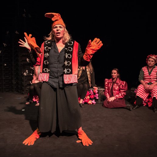 Agnieszka stoi na środku sceny, w czarno-pomarańczowym kostiumie, na dłoniach ma koślawo założone rękawiczki, czapka ma również rękawiczkę. Mówi w ożywieniu.