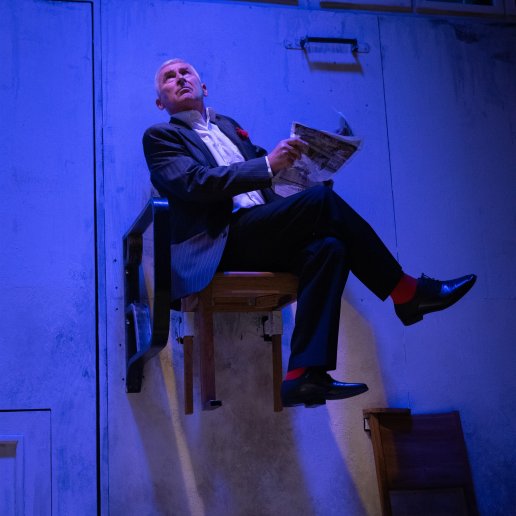 Elegancki aktor w garniturze i białej koszuli siedzi na krześle i trzyma gazetę. Krzesło zamocowane jest na ścianie, wisi nad podłogą.