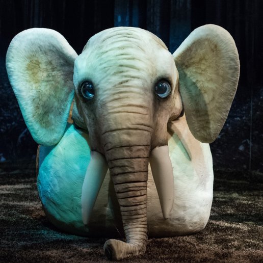 Wielka lalka słonia. Słoń ogromny, ma duże szkliste oczy, siedzi na ziemi w lesie. Obok niego aktorka, trzyma w ręce lalkę myszy.